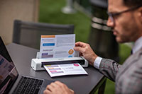 Homme portant une veste grise avec des lunettes, assis à l'extérieur en utilisant un ordinateur portable et le scanner de documents portable DS-740D de Brother scannant un document A4 en couleur