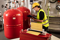 Ingénieur portant un casque jaune et une veste haute visibilité à genoux et prenant des notes, avec le scanner de documents portable DSmobile DS-940DW de Brother dans la salle des machines