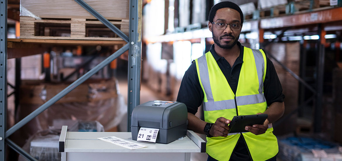 Un employé d'entrepôt portant un gilet à haute visibilité tient une tablette à côté d'une imprimante de bureau Brother TD qui imprime des étiquettes.