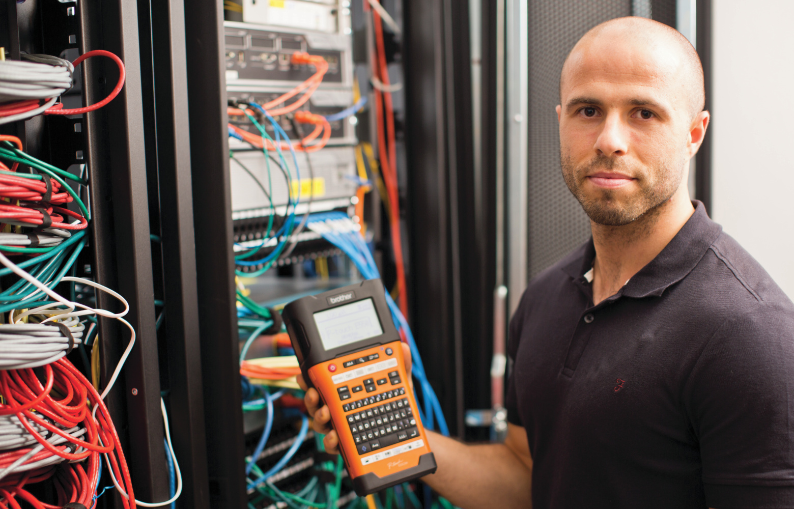 Un technicien réseau se tient devant une armoire réseau soigneusement étiquetée, tout en tenant une imprimante d'étiquettes P-touch