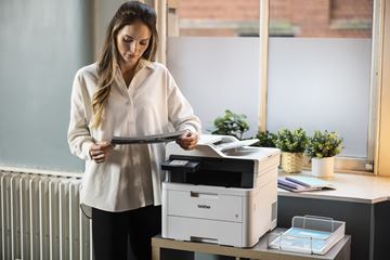 Une femme est installée devant une imprimante laser Brother. Elle tient dans sa main des documents quelle vient d'imprimer.