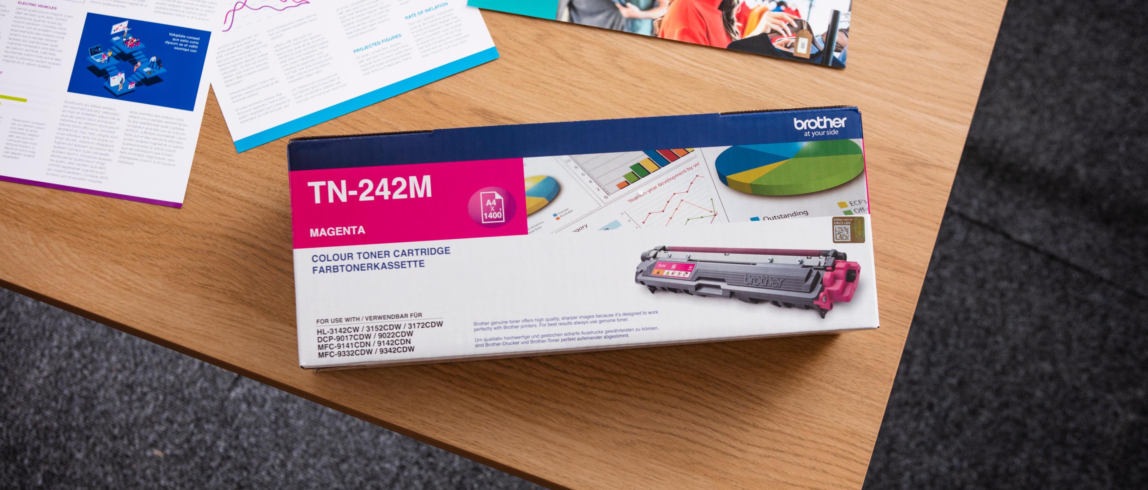 A top shot of a boxed Brother toner cartridge on a light brown office table against a dark grey carpet background