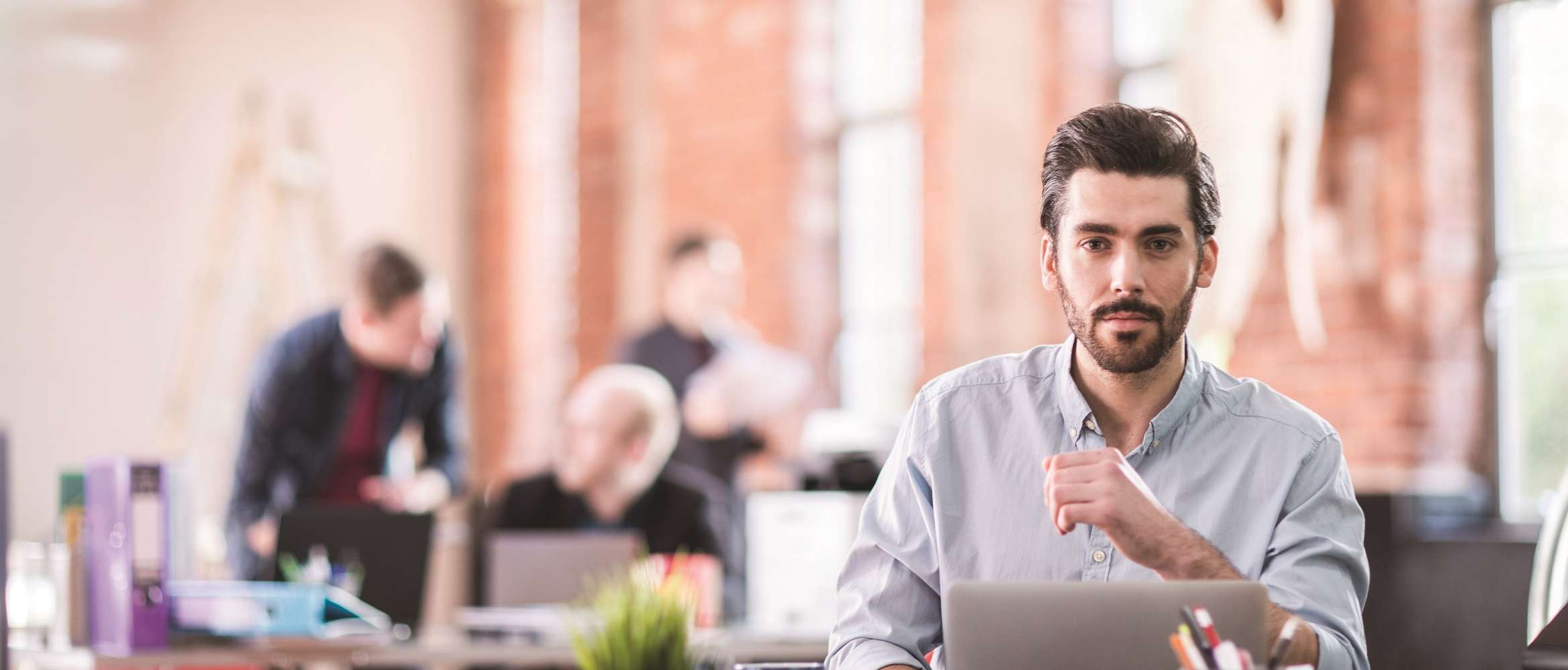 A man dressed in smart casual attire is sat working at a laptop with three co-workers in the background of the casual office space