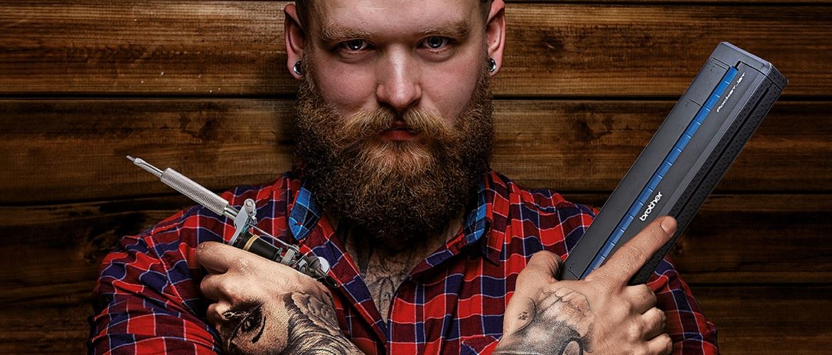 Uomo con barba e camicia a quadri con in mano Brother Stencil Machine e ago per tatuaggi