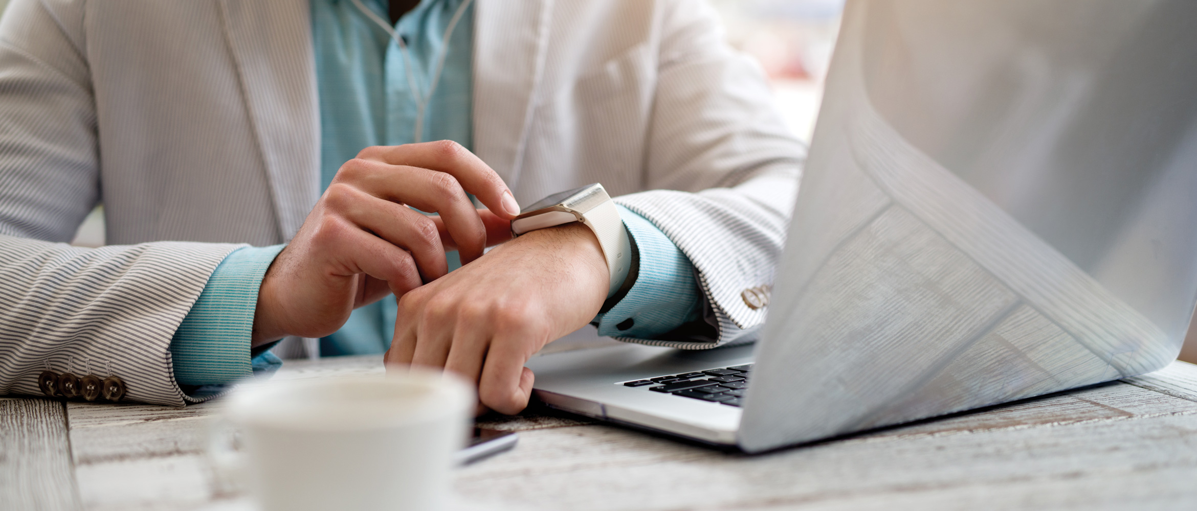 A man sitting next to his office laptop is wearing a grey suit and blue shirt. He checks his wearable technology smartwatch.