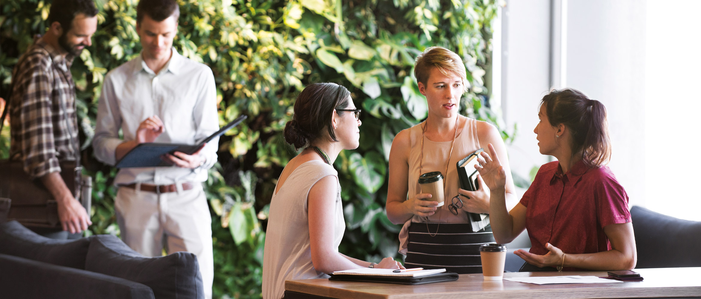Two men and three women are holding an informal business meeting in the workplace of the future that features a prominent living green wall of plants within the office environment.