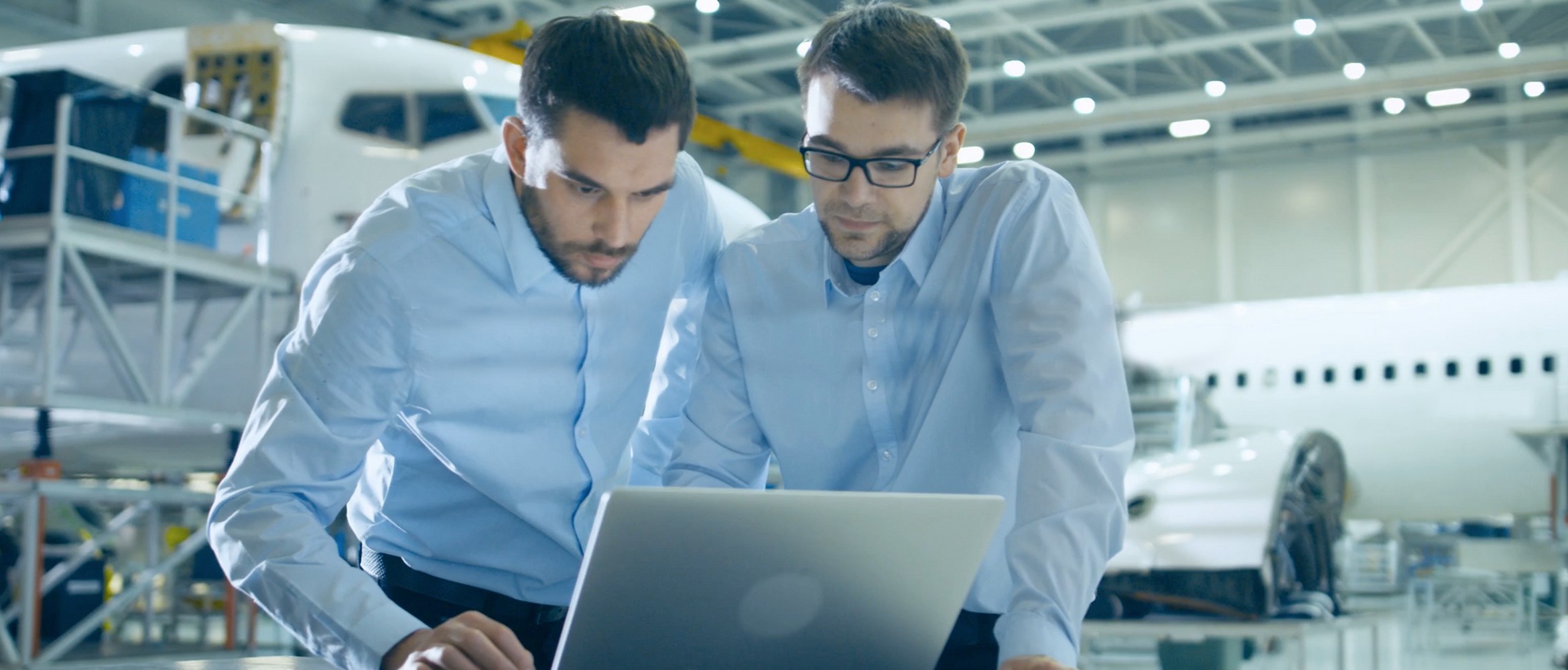 Two business men are remotely working outside of the office. They are meeting around a laptop set up in an aircraft hanger.