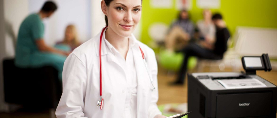 A female doctor is wearing a traditional white medical coat with a stethoscope hanging around her neck. She's standing in a hospital waiting room with patients and healthcare staff in the background. She is standing next to a Brother multi-function business printer. 