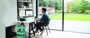 A male staff member is working from a home office / hybrid working. He is sat at a desk working on a desktop computer with a Brother P-touch labelling printer at his side to increase business productivity.