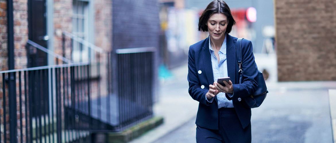 A woman wearing a business suit is walking through a city residential street while checking her smart phone for updates