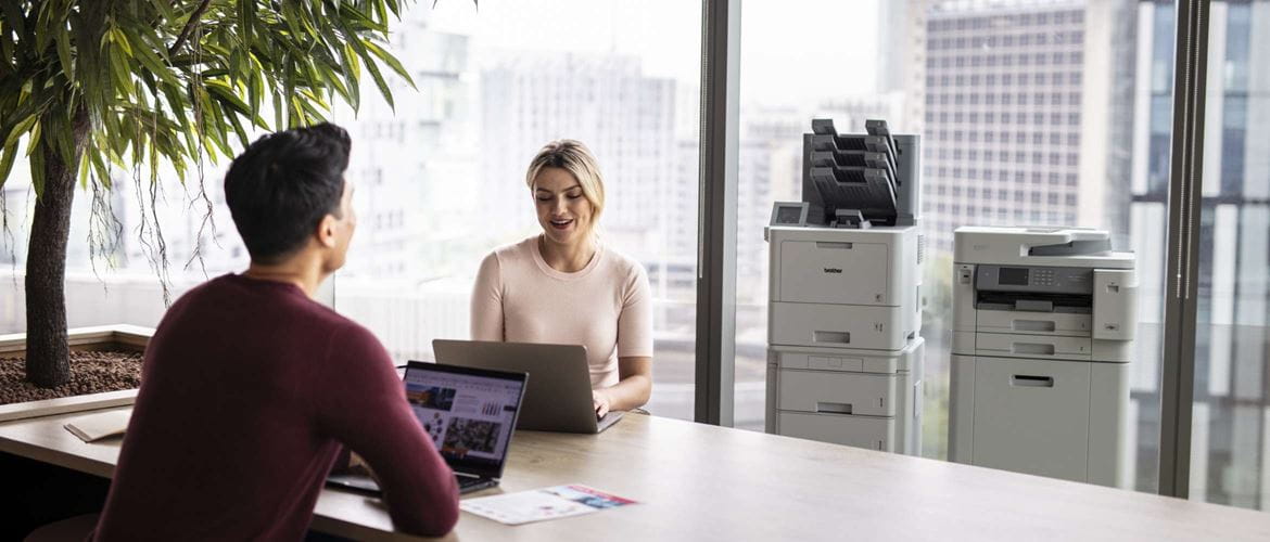 Two office workers in a brightly lit business space are working at a desk with two Brother floor-standing SMB printers and a city skyline in the background