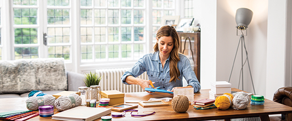 Woman using scissors sat at a table covered with boxes, jars, balls of wool and folders