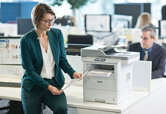 Woman wearing glasses, green suit sat on table next to Brother MFC-L6900DW printer, man wearing suite, monitors, desks
