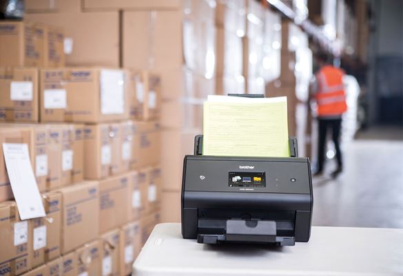 Brother ADS-3600W with dispatch notes in warehouse, warehouse worker in orange hi-vis in background, boxes, table