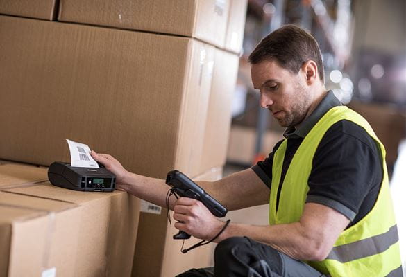 Man wearing hi-vis holding scanner printing label, label printer on top of boxes in warehouse
