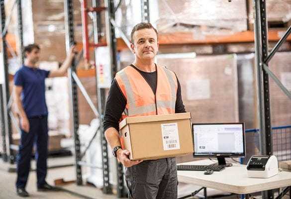 Man with short brown hair in orange hi-vis holding brown box in warehouse, table, monitor, keyboard, mouse, racking