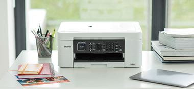 Brother DCP-J572DW inkjet printer on white desk, laptop, pen pot with pens, note books