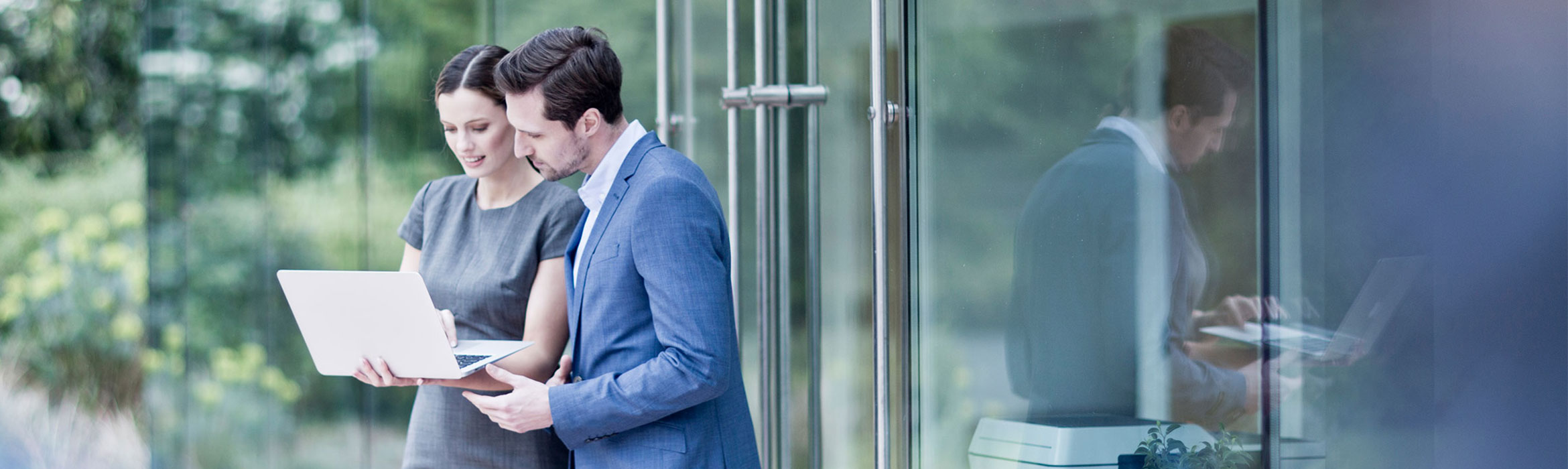 Man with dark hair wearing blue suite white shirt stood speaking to woman wearing grey dress holding laptop outdoors, glass door, printer inside, green bushes