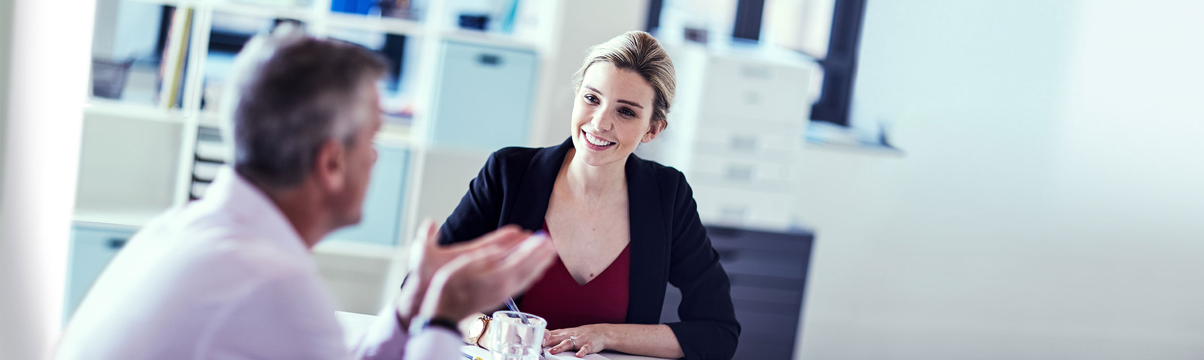 Female with hair tied back wearing black jacket red top sat speaking to man wearing light pink shirt, pen, glass, paper, Brother printer, shelf, box