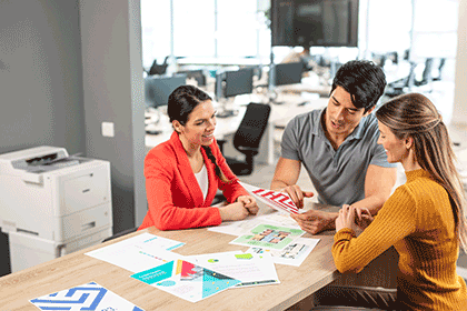 A man and two women sat around table with colour documents on the table, printer, desks, chairs