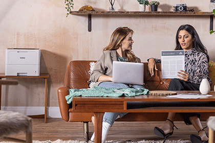 Two woman sat on sofa talking with laptop, mono A4 document, Brother HL-L6210DW, wooden table