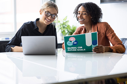 Two women sat next to each other, one holding colour A4 document, laptop