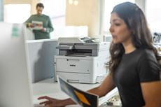 Female sat at desk facing monitor, holding colour document, Brother MFC-L8340CDW printer, male holding colour document