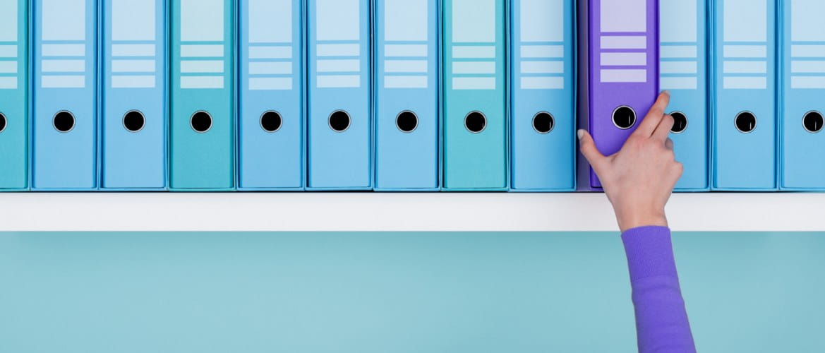 A shelf full of similar looking teal blue-green lever arch files has one purple file folder standing out. It is being picked up by an office staff member wearing a matching purple item of clothing. 