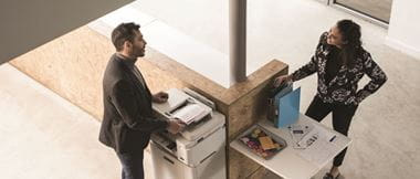 A man and a woman stop in a brightly lit office to talk by a business printer and some files