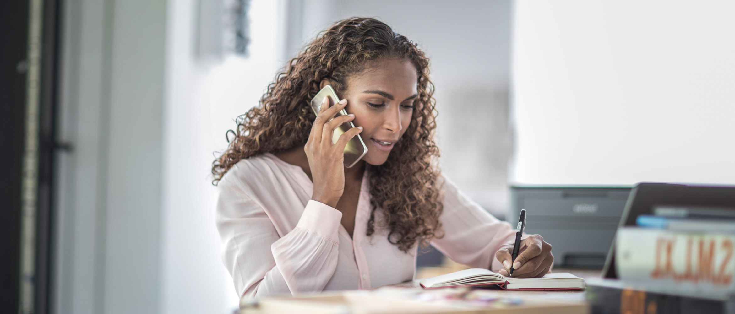 Professionally dressed woman at her desk speaking on her mobile phone and taking notes with Brother printer in background