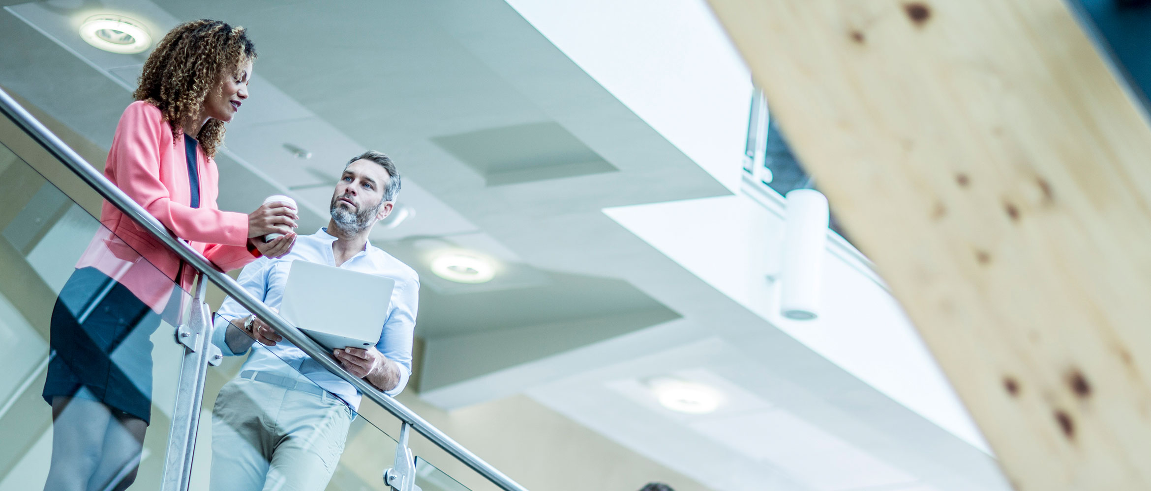 A man holding a laptop listens to a woman colleague talking, stood on balcony