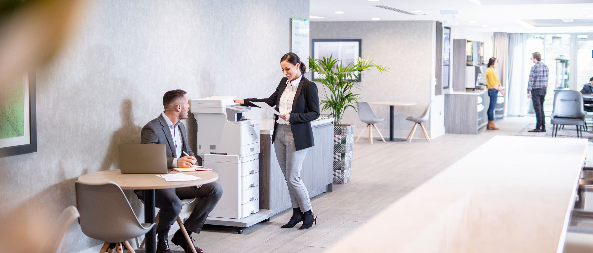 Woman wearing suit stood at Brother MFC-L9570CDWTT all in one colour laser printer, man in suite sat at table with laptop, plant, people at the back