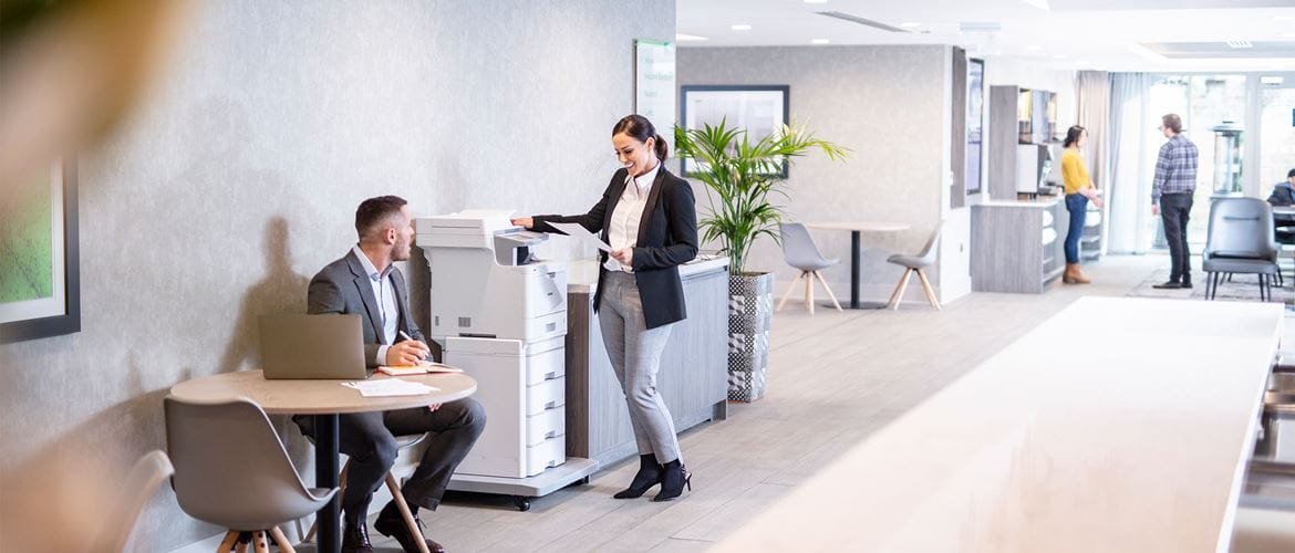 Woman wearing suit stood at Brother MFC-L9570CDWTT all in one colour laser printer, man in suite sat at table with laptop, plant, people at the back
