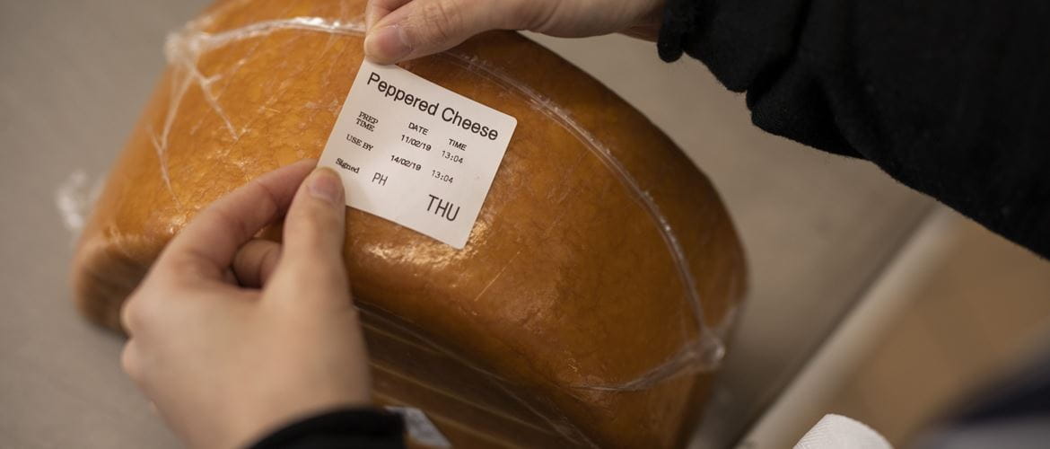 A member of a kitchen staff team applies a food information day label onto a block of peppered cheese