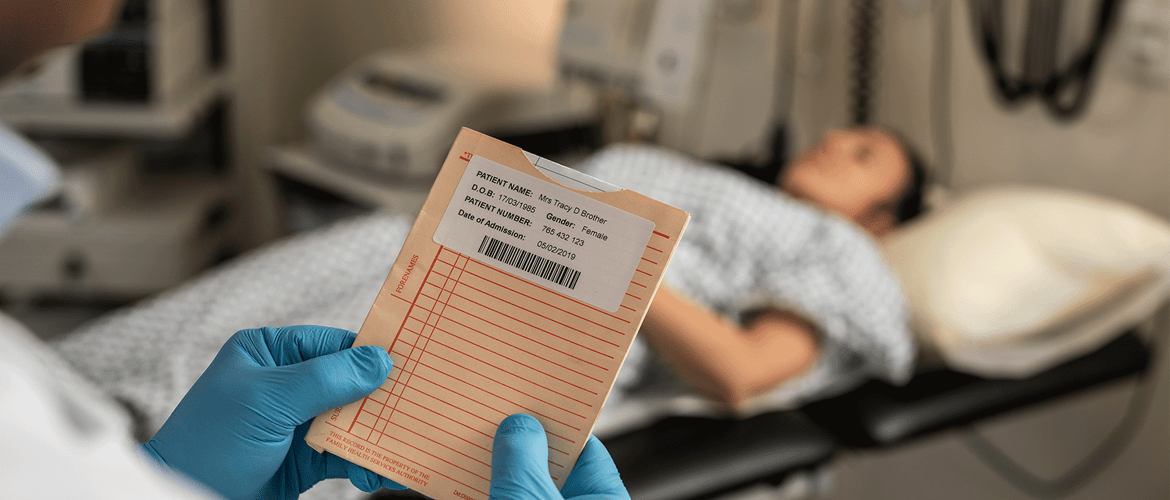 En sykehusansatt holder et dokument med en etikett med strekkoder, og en pasient ligger på et undersøkelsesbord i bakgrunnen