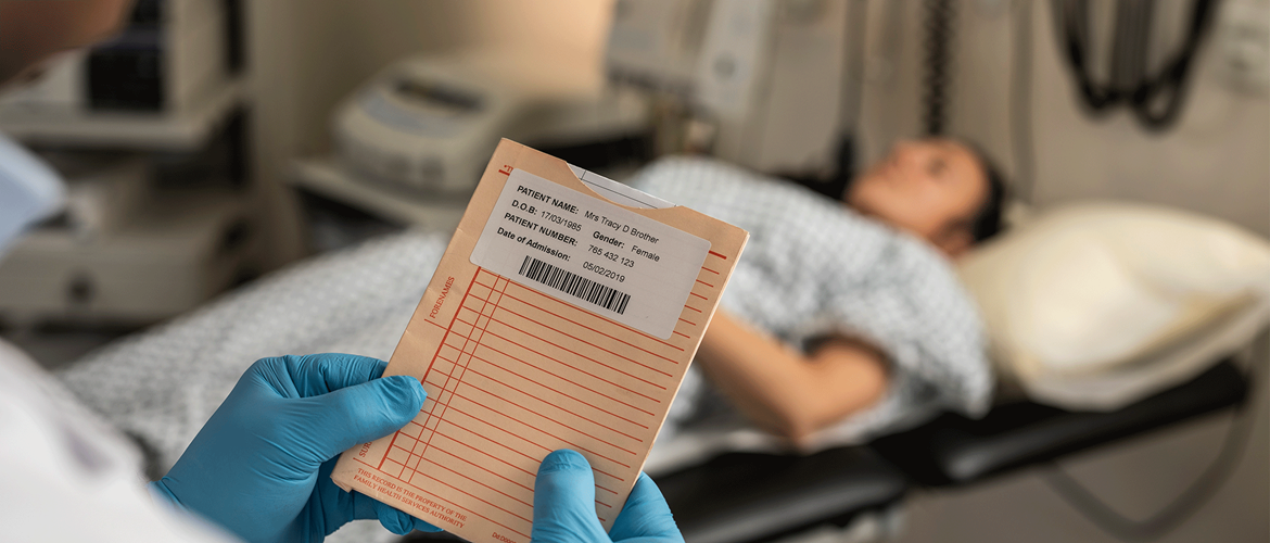 En sykehusansatt holder et dokument med en etikett med strekkoder, og en pasient ligger på et undersøkelsesbord i bakgrunnen