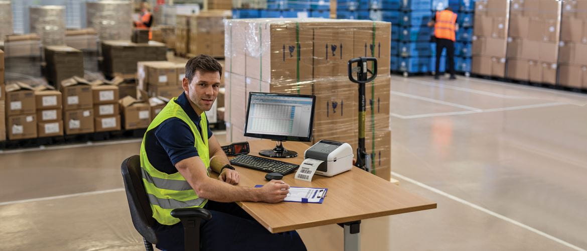 Man sat in chair at desk wearing yellow hi-vis in warehouse with monitor, keyboard, Brother TD label printer, boxes, pallet truck