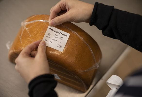 A member of a kitchen staff team applies a food information day label onto a block of peppered cheese