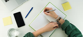 En kvinne skriver i en notatbok, en mobiltelefon, et glass vann, en  penn og post-it blokker ligger på skrivebordet