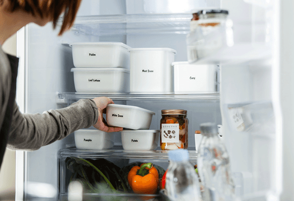 Nainen laittaa jääkaappiin valkoisia ruoka-astioita, jotka on merkitty musta-valkoisilla teksteillä