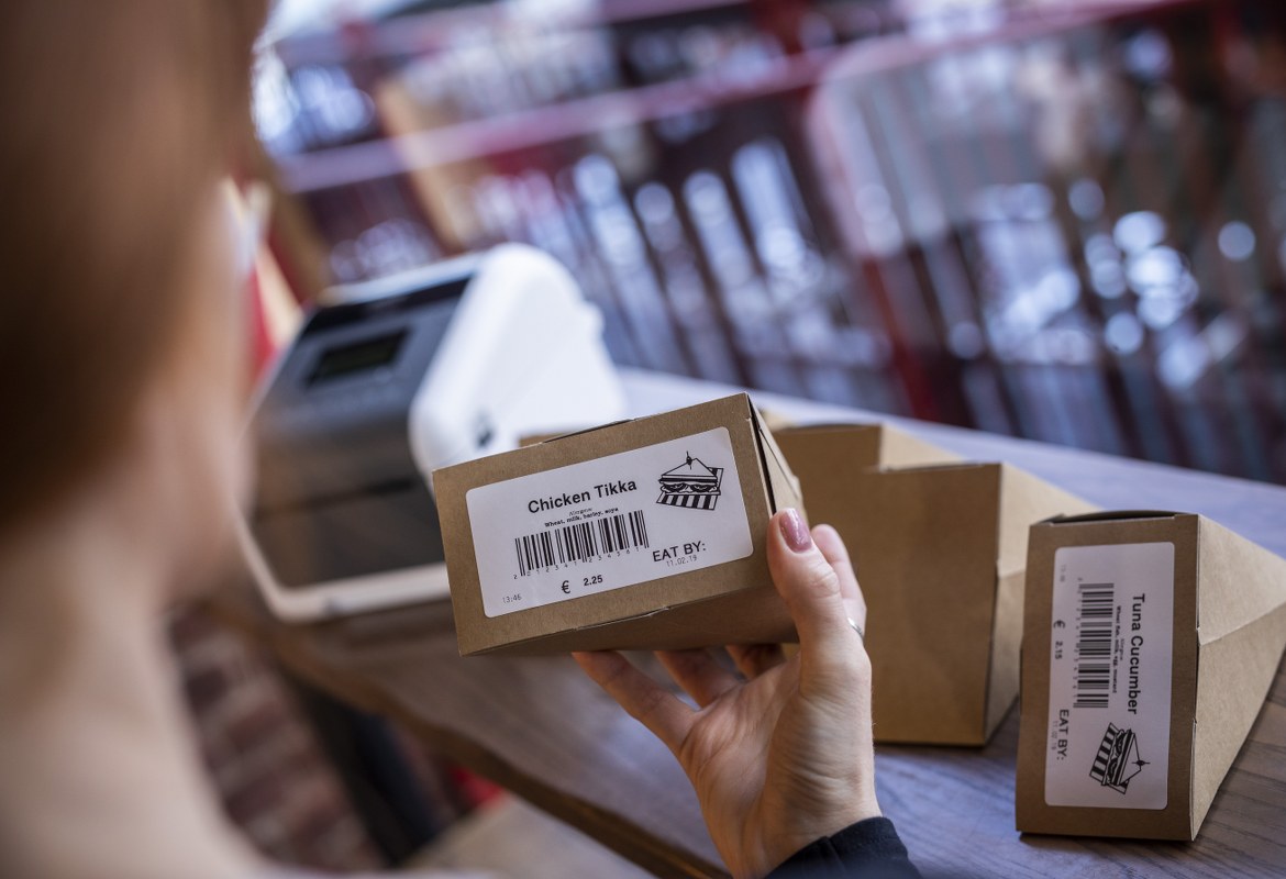 A female member is checking the printed food labels from a Brother labelling machine that have been added to prepacked sandwiches in a food deli or bakery shop store environment. They labels contain a barcode, title, image, price and use by date. 