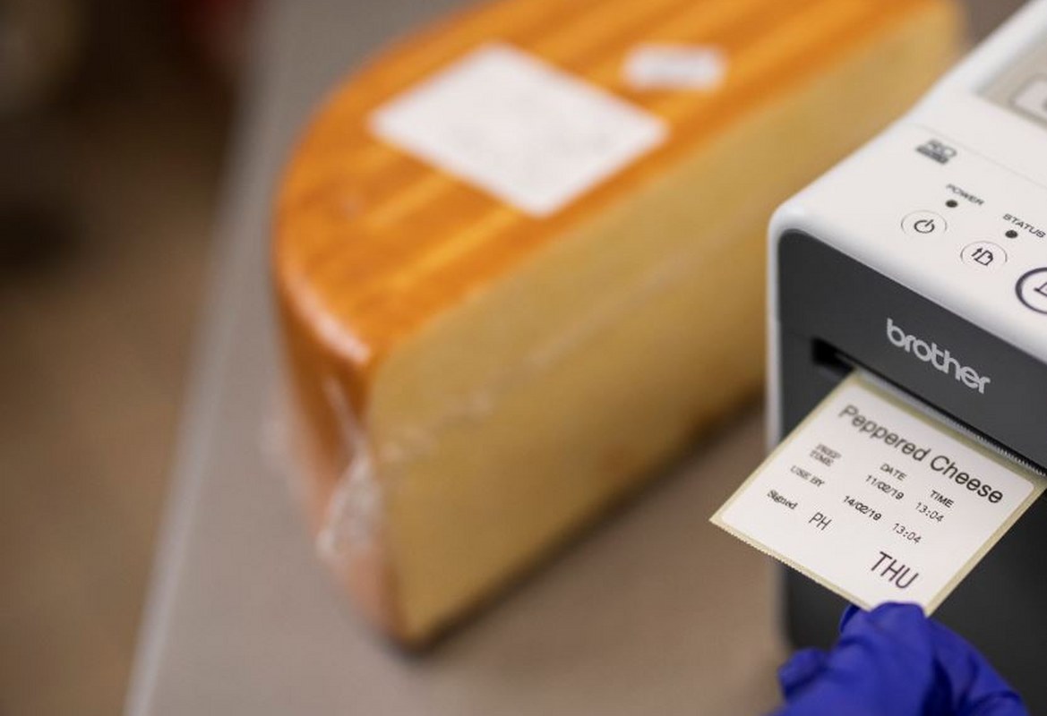 Close up a Brother label printer printing out a food day label for a block of cheese in the back office of a food deli or cafe