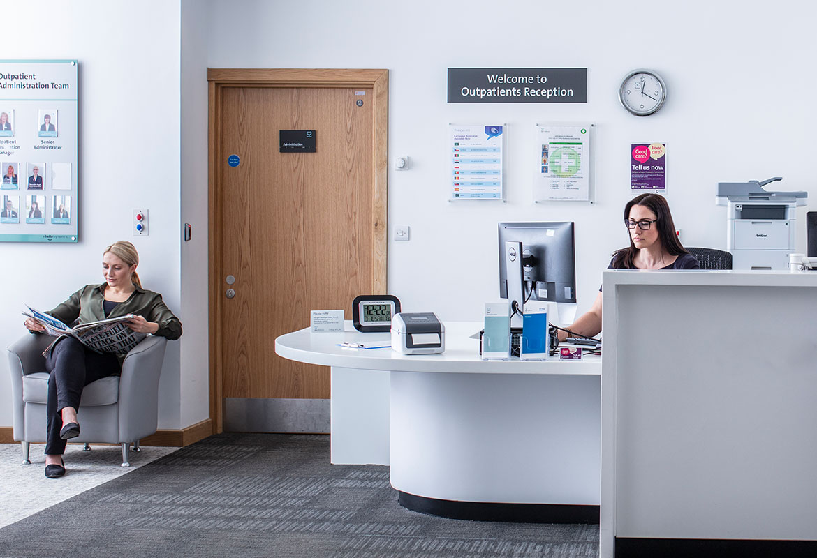 Female receptionist sat at desk using a computer, female patient sat reading newspaper
