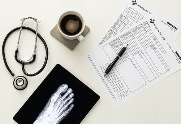 Medical forms, pen, cup of coffee on coaster, stethoscope, tablet with xray of foot on white table