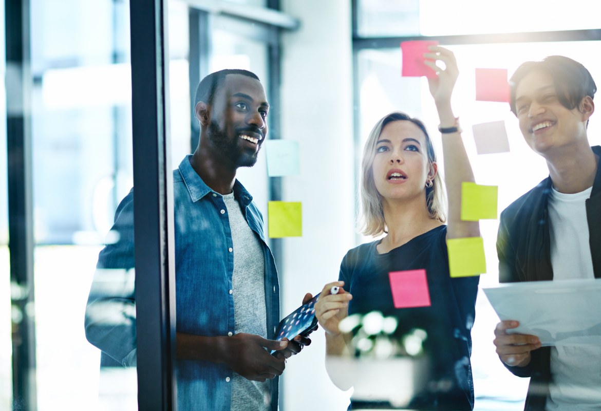 Three office workers stick post-it note stickers onto a glass wall in a moment of creative collaboration during a business meeting
