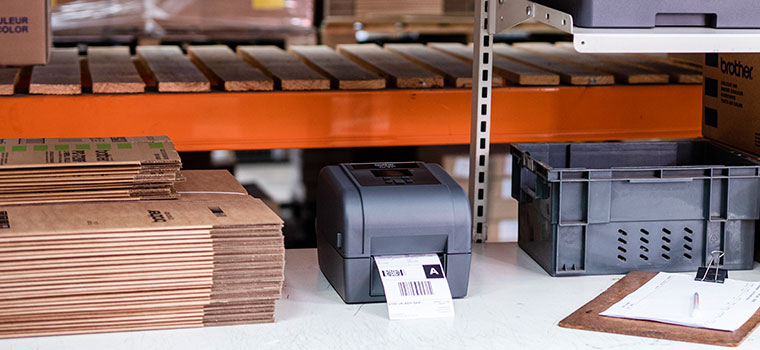 Brother grey label printer on desk, brown boxes, clipboard, grey crate, orange racking