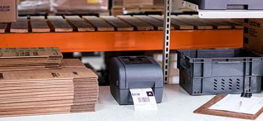 Brother grey label printer on desk, brown boxes, clipboard, grey crate, orange racking