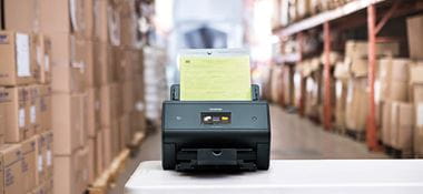 Brother ADS-3600W desktop scanner on table in warehouse with boxes on racks