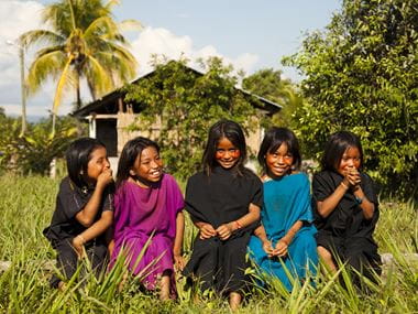 Fem jenter i fargerike klær sitter i gresset utenfor et hus i en landsby