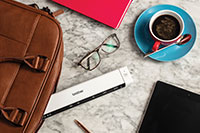 Pöydällä silmälasit, kahvikuppi, kynä, mobiiliskanneri, tabletti, pinkki muistikirja ja nahkainen läppärilaukku